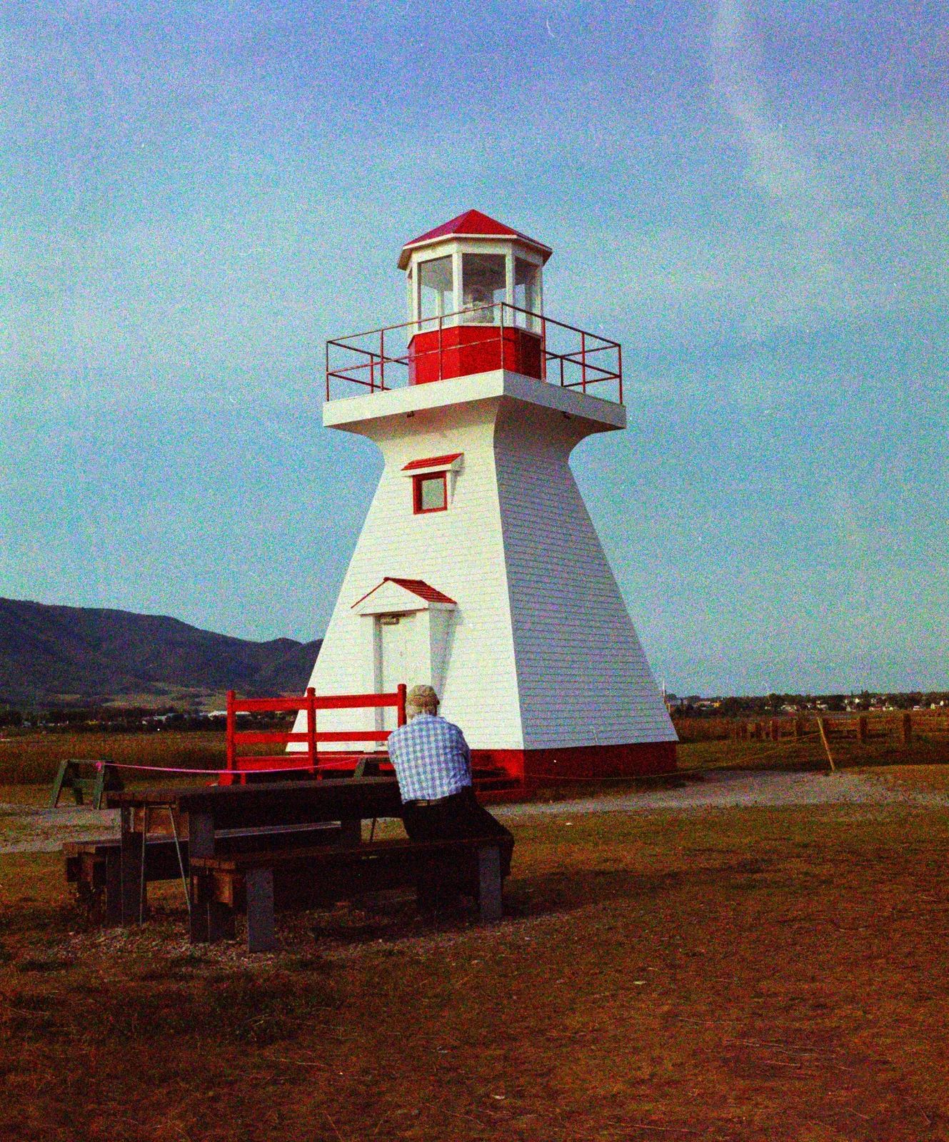 A red and white lighthouse, with a man on a bench below, converted with Sunny 100